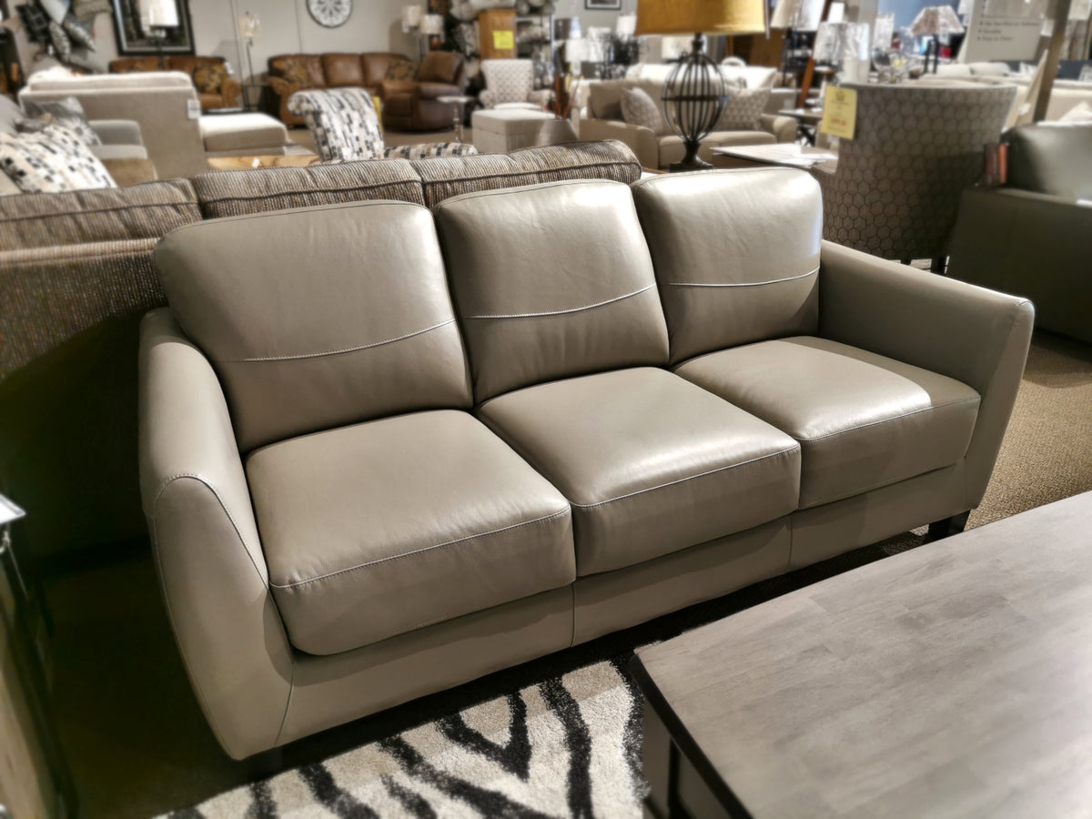 The Leather Craft Oxford Sofa in gray leather with three seat cushions sits beside a wooden coffee table in the showroom.