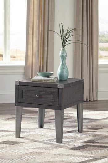 The Ashley Tadoe End Table with a drawer, holding a vase, bowl, and books, is set on a patterned rug by large windows.