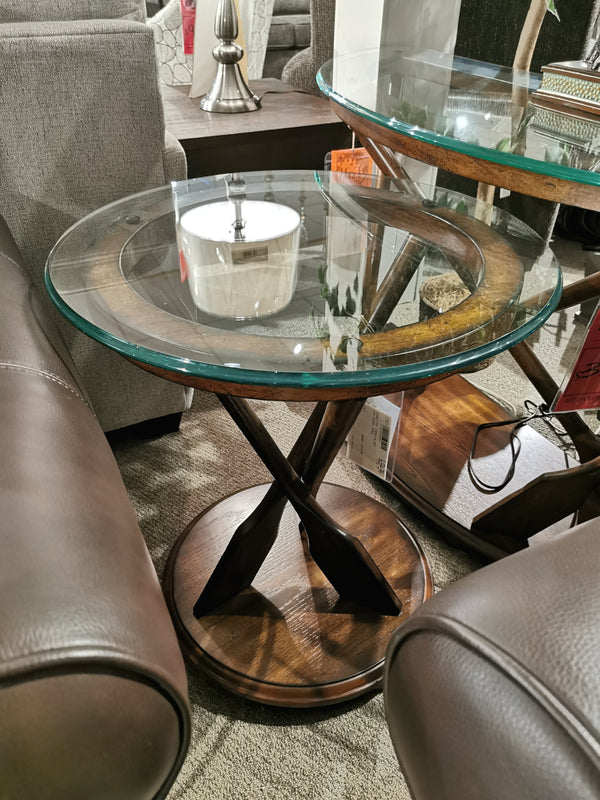 The T2214 Beaufort Round End Table by Magnussen sits between brown leather sofas in a showroom.