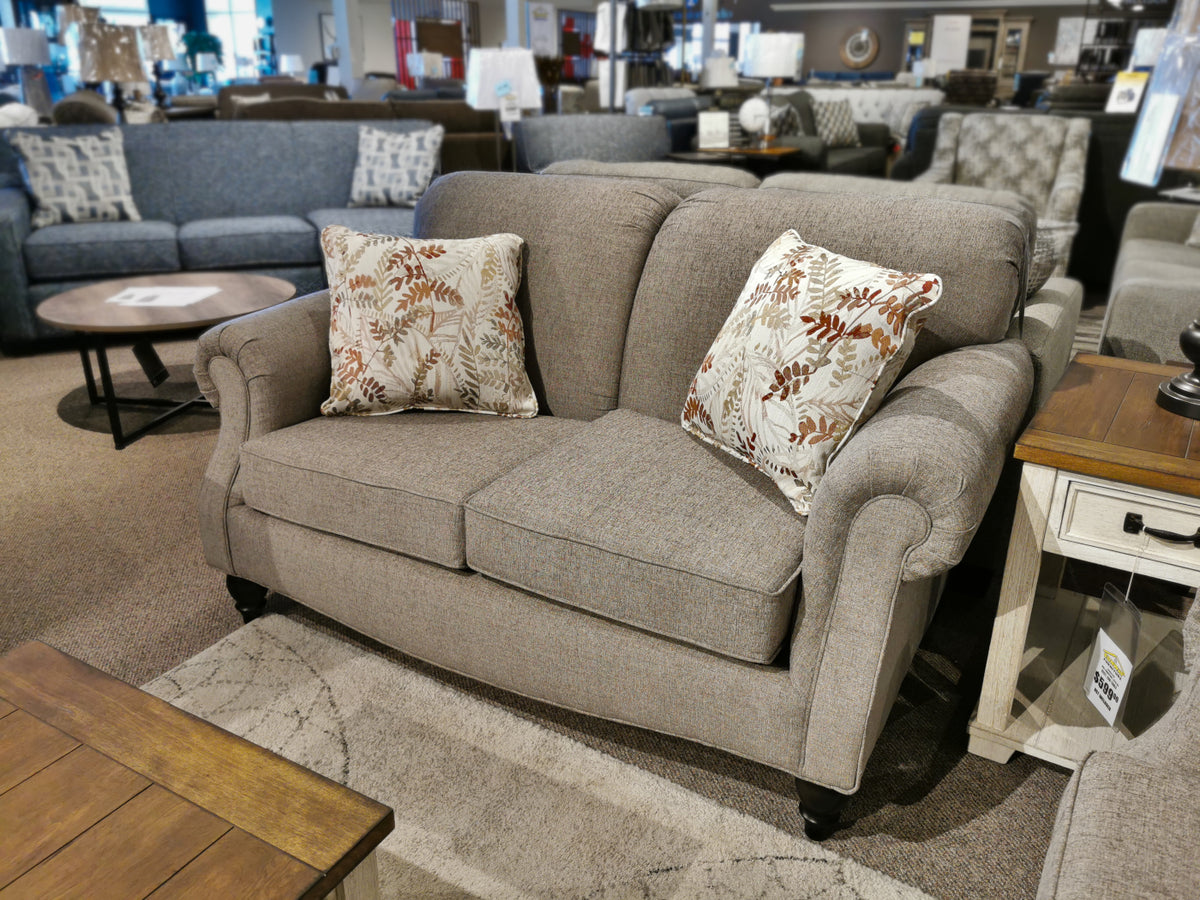 The Superstyle 7303 Loveseat is a gray Canadian sofa with rolled arms and two patterned pillows, shown in a showroom.