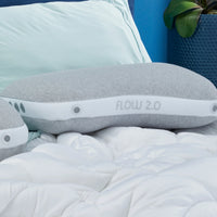 A gray Bed Gear Flow Cuddle Pillow rests on a white comforter with light blue sheets and a potted plant nearby.