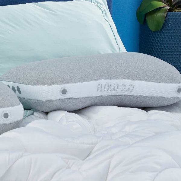 A gray Bed Gear Flow Cuddle Pillow rests on a white comforter with light blue sheets and a potted plant nearby.