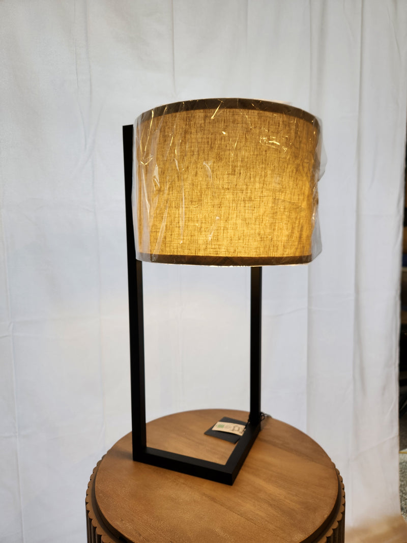 The Thorton Black Table Lamp by Pacific Coast features a sleek black metal frame and beige shade on a round wooden table.
