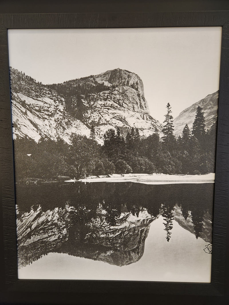 Black and white photograph titled "15291 Mirro Lake" by Mercana, featuring a mountainous backdrop and trees reflected in the serene water, elegantly framed in a dark wooden frame.