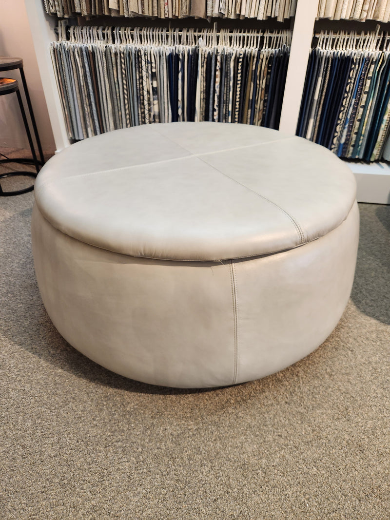 The 301 Oliver Round Storage Ottoman by La-z-boy has visible seams, enhancing a room with fabric swatches on vertical shelves.