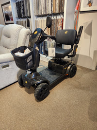 The Golden Companion Full-Size Mobility Scooter, black with 4 wheels and a front basket, is shown indoors on carpet.