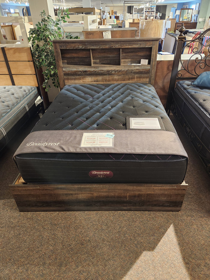 A Beauty-rest ESME TT Medium queen mattress on a rustic bed frame with bookshelf headboard in a furniture store.