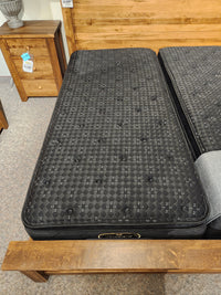 A black Beauty-rest Adonis PT Plush twin mattress rests on a wood bed frame beside a matching nightstand.