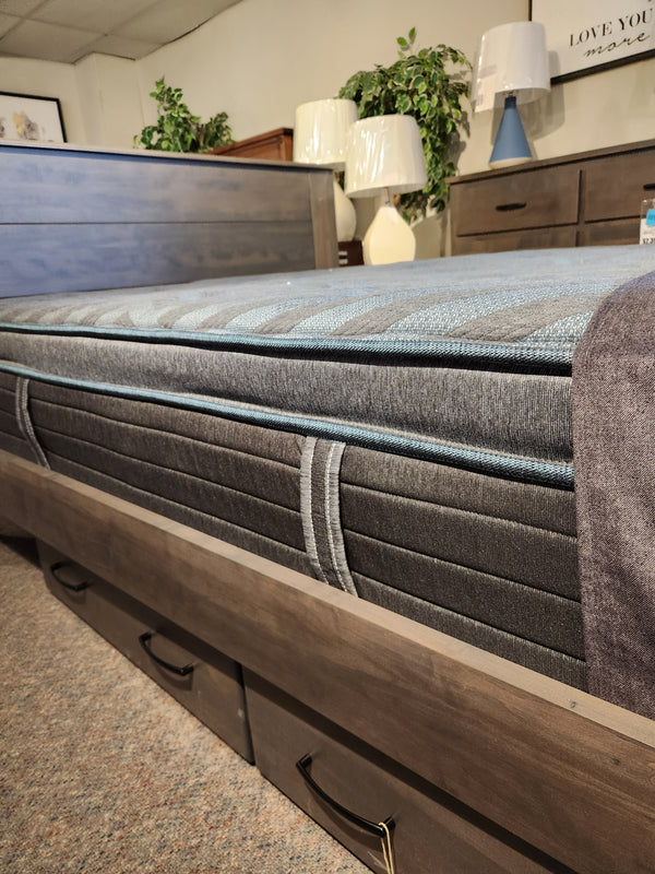 A close-up of the Beauty-rest Isla PT Firm Mattress on a wooden bed frame, with lamps and plants in the background.