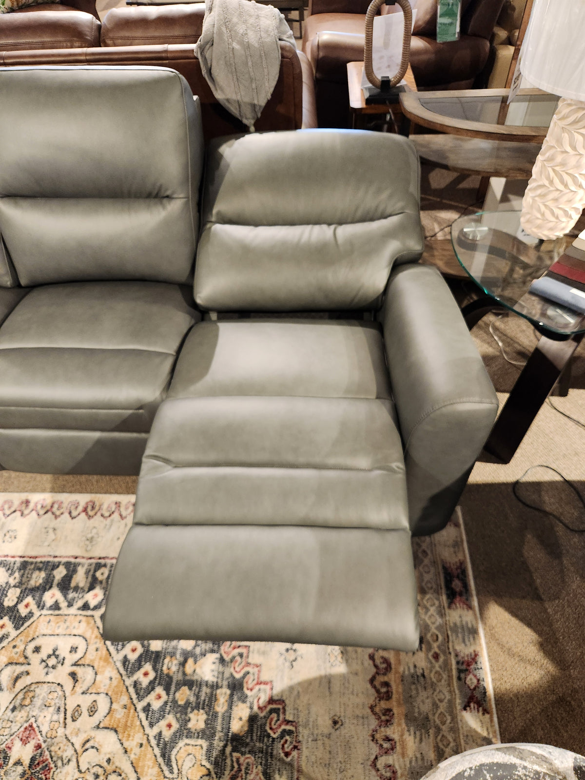 A Leather Craft Texas Power Reclining Loveseat with side table and lamp sits on a patterned rug in a showroom.