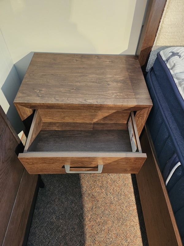 The Magnussen Llewellyn B5907 Night Stand with a walnut finish and single drawer is beside a bed with a blue mattress.