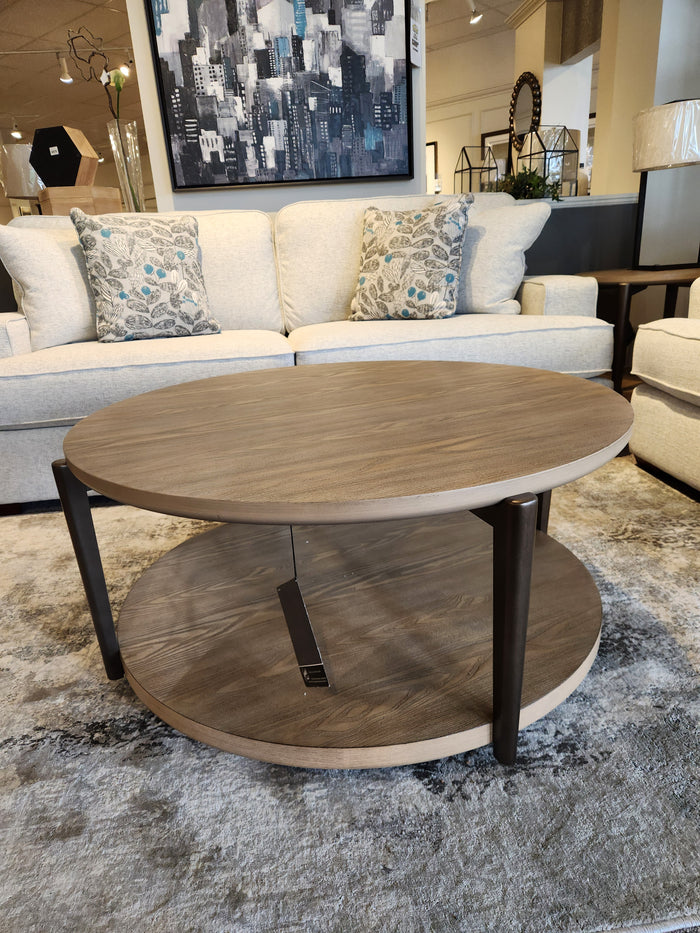 The Ashley Dyonton Round Cocktail Table features a wood-tone finish and lower shelf for an industrial, modern showroom look.