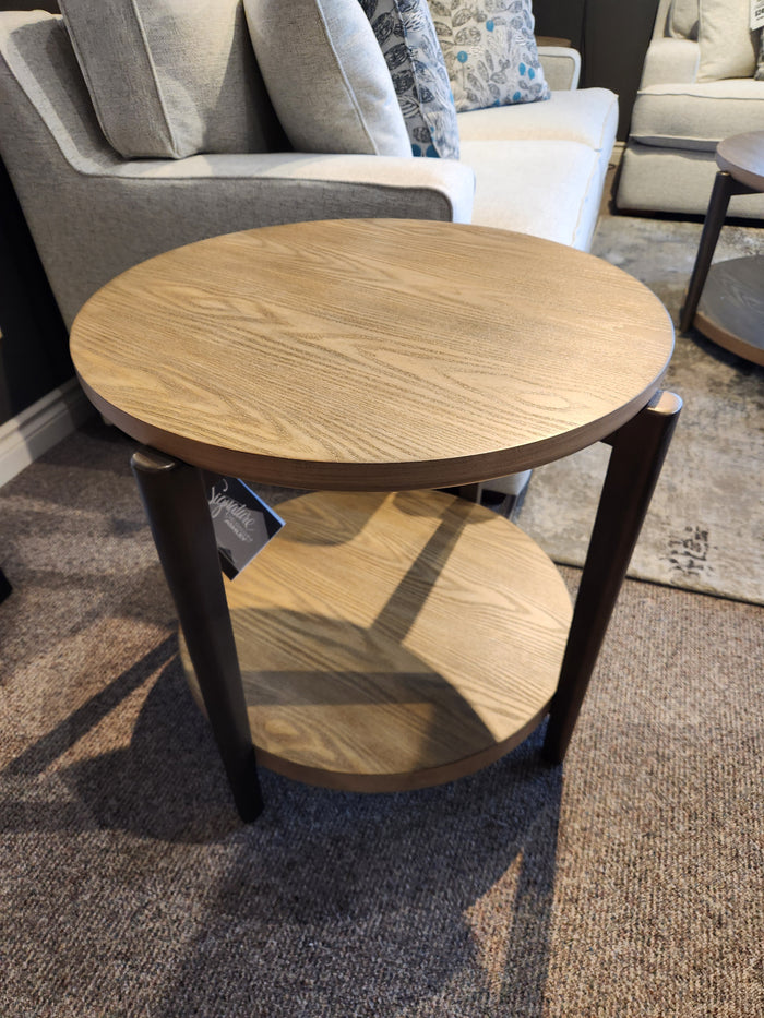 The Ashley Dyonton Round End Table with wood-tone finish and shelf sits on carpet next to a light gray sofa.