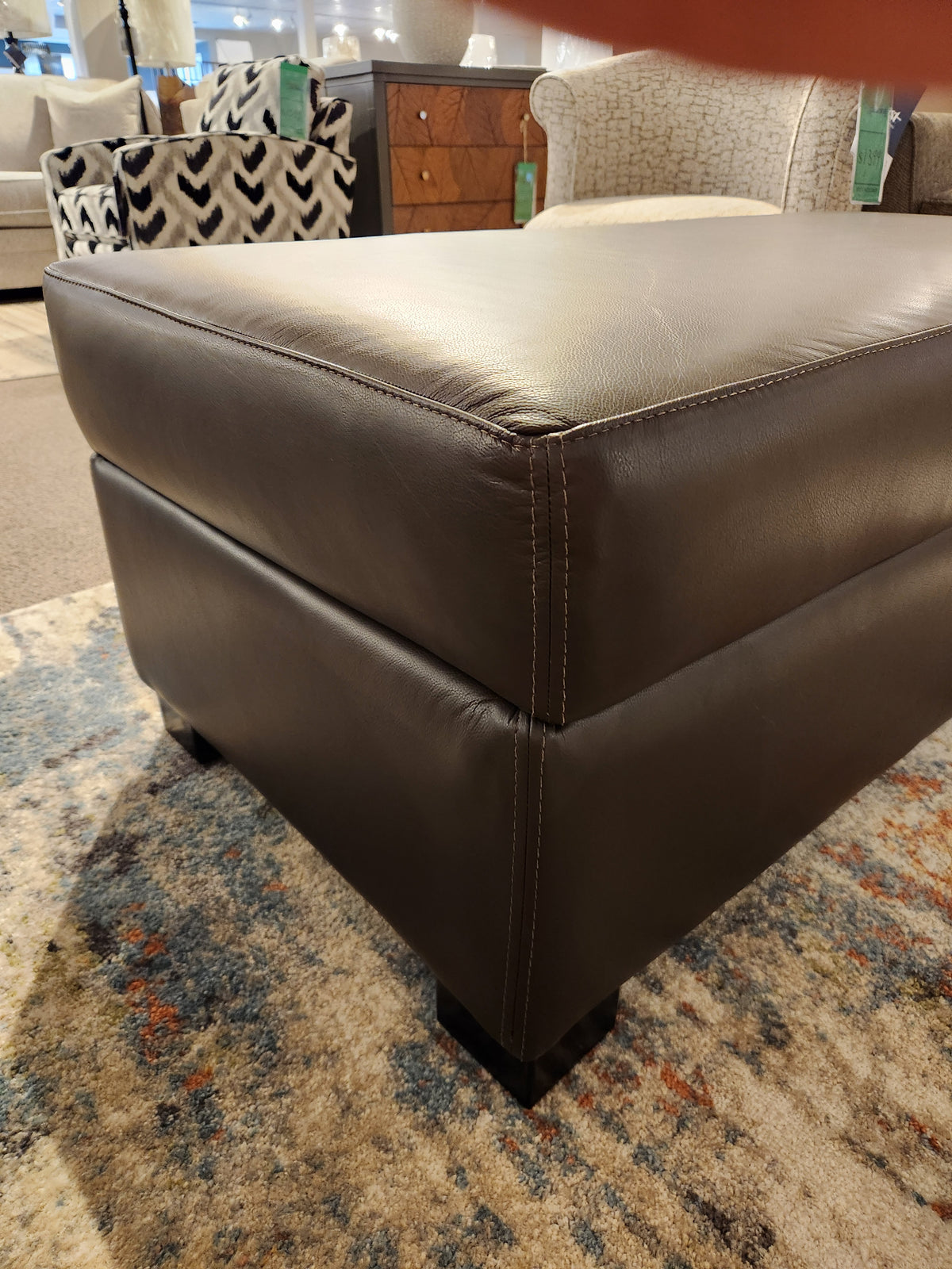 Close-up of a Superstyle 5000-57 Storage Ottoman with visible stitching, placed on a patterned rug in a showroom.