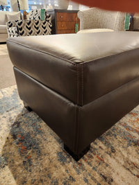 Close-up of a Superstyle 5000-57 Storage Ottoman with visible stitching, placed on a patterned rug in a showroom.