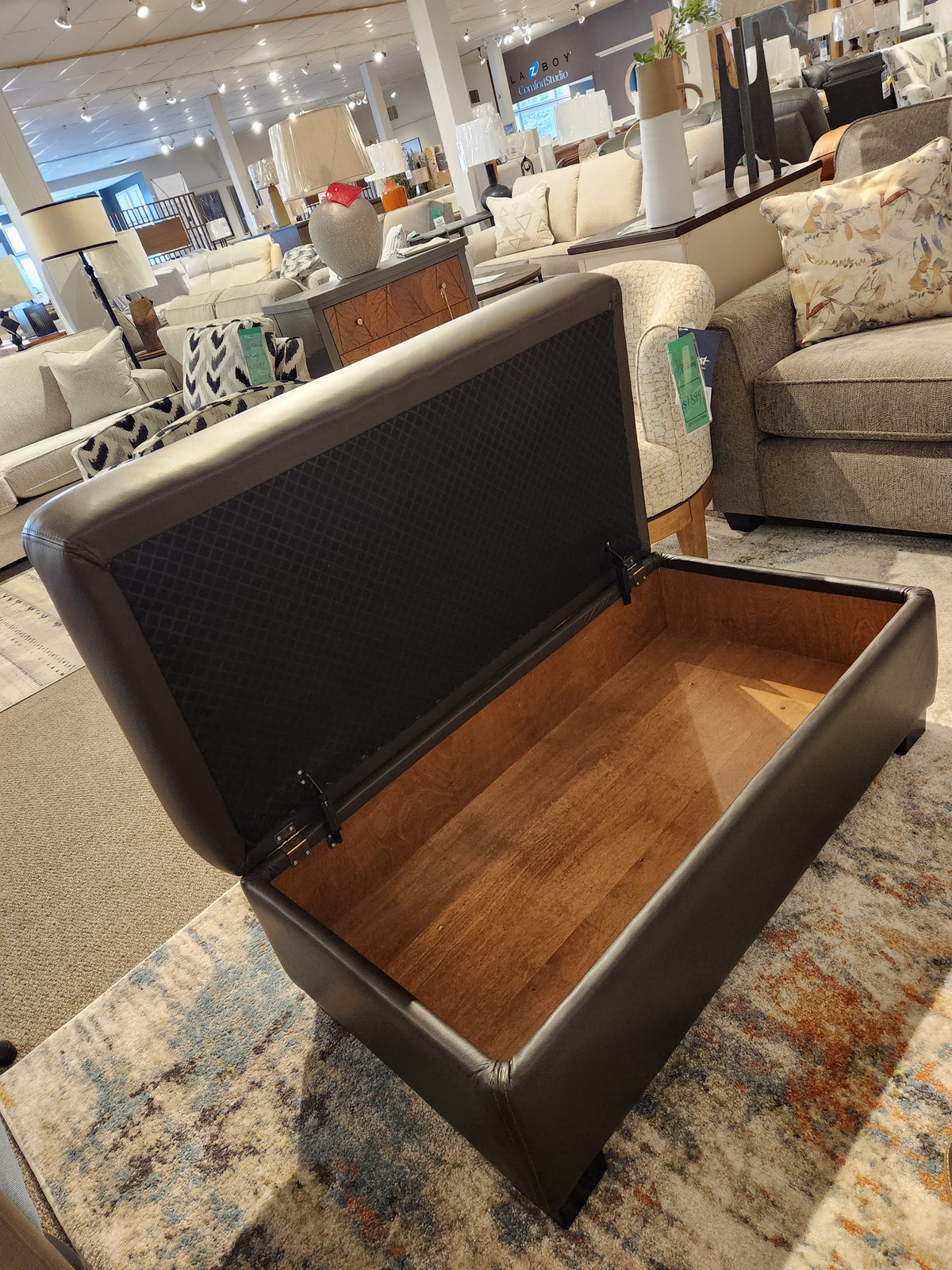 A dark brown Superstyle 5000-57 Superstyle Storage Ottoman with its lid open, displayed in a furniture showroom.
