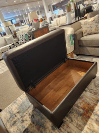 A dark brown Superstyle 5000-57 Superstyle Storage Ottoman with its lid open, displayed in a furniture showroom.