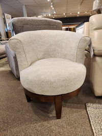 The Attica Swivel Chair by Best features beige upholstery, a curved back, and wood base, shown in a furniture showroom.