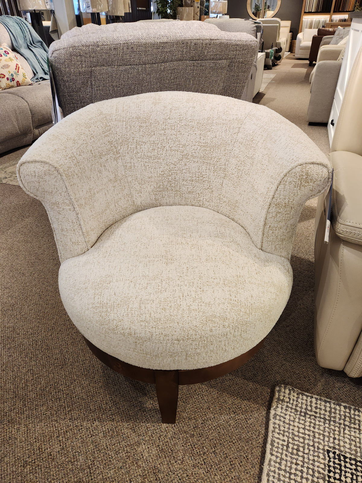 The Attica Swivel Chair by Best features curved arms and wood legs in beige, showcased in a furniture showroom.