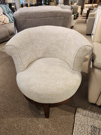 The Attica Swivel Chair by Best features curved arms and wood legs in beige, showcased in a furniture showroom.