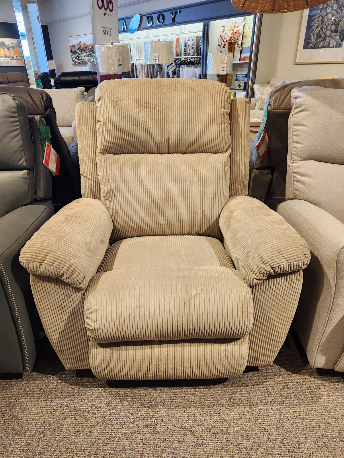 The La-z-boy 761 Joel Reclining Chair in beige with cushioned arms is shown among recliners in a showroom.