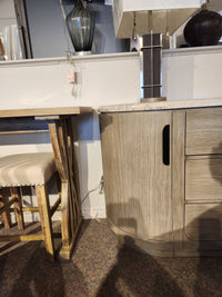 Magnussen D5874 Braelyn Buffet w/Stone Top stands beside an upholstered chair, lamp, and wooden table on carpet.