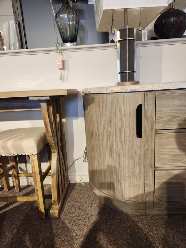 Magnussen D5874 Braelyn Buffet w/Stone Top stands beside an upholstered chair, lamp, and wooden table on carpet.