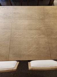 Magnussen D5878 Trestle 5 Piece Dining Package in Honey Wheat with two white-cushioned chairs partially visible.