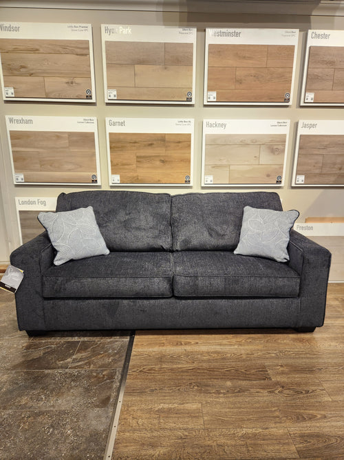 An Ashley Altari Queen Sofa Bed in dark gray with a foam mattress and patterned pillows sits by wood flooring samples.