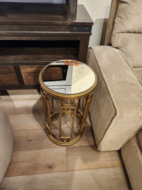 The Magnussen T6300 Lorena round accent table with mirrored top sits between a beige sofa and TV stand on hardwood floor.