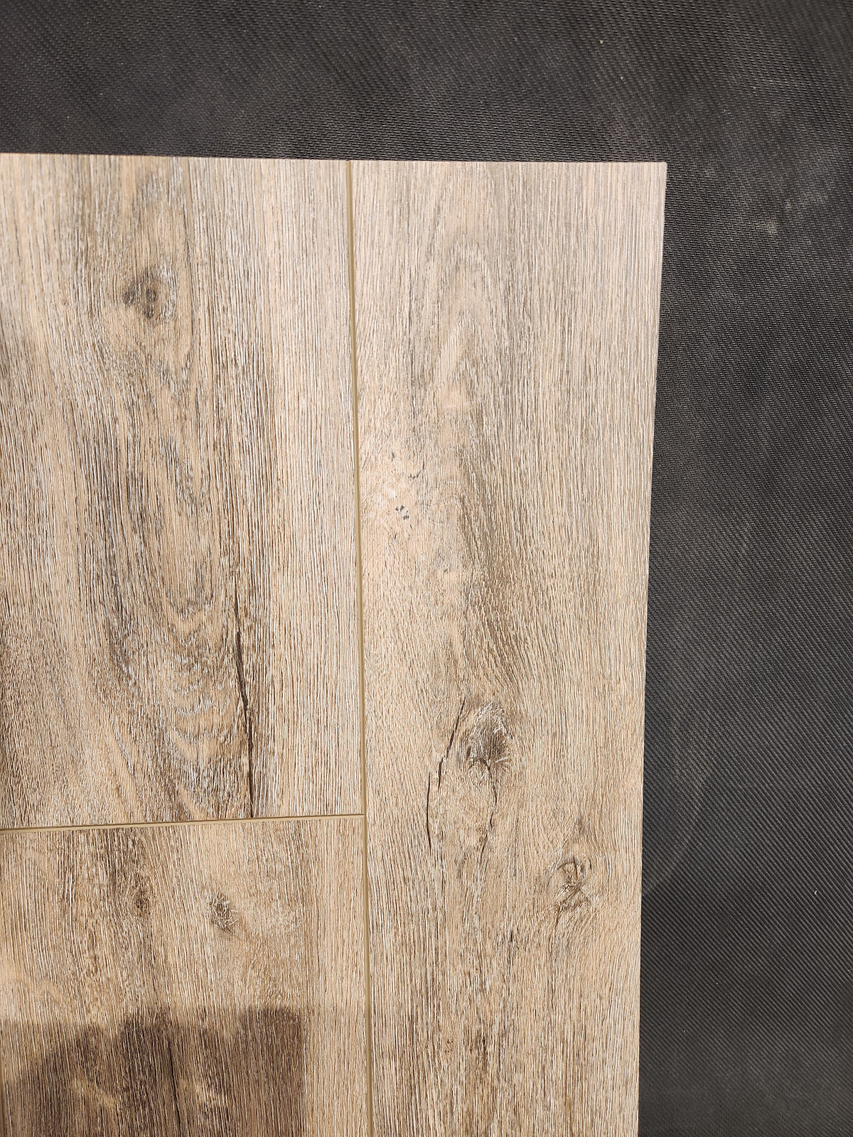 Hang Loose Luxury Vinyl (Glue-Down Plank) by Beckham Brothers, with a light brown grain, is laid on a dark textured surface.
