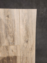 Hang Loose Luxury Vinyl (Glue-Down Plank) by Beckham Brothers, with a light brown grain, is laid on a dark textured surface.