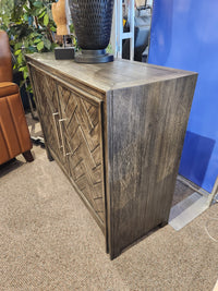 The Jofran Gramercy 2 Door Accent Cabinet features chevron inlay and metal handles, styled near plants and furniture.