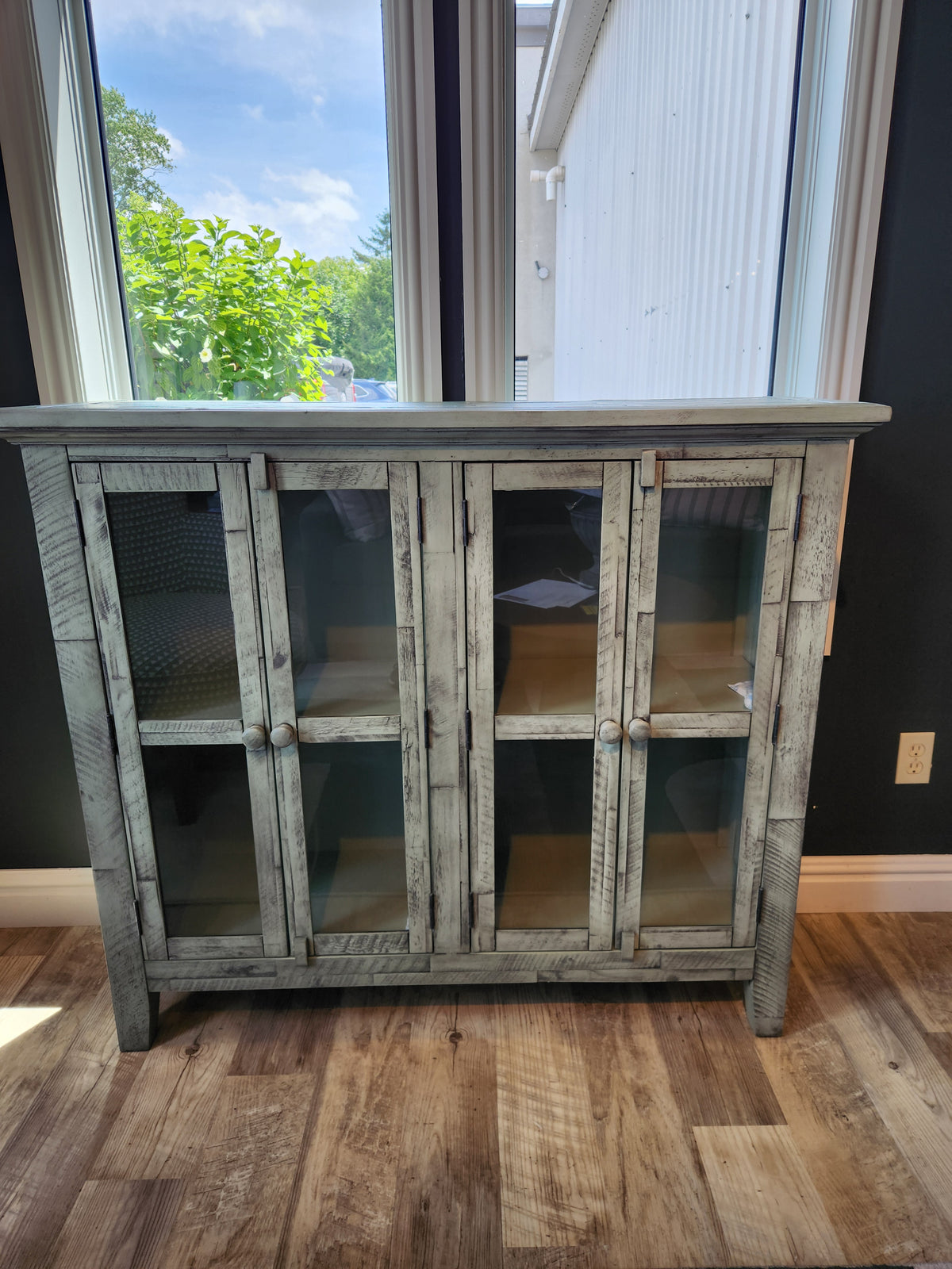 The Jofran Rustic Shores 4 Door Accent Cabinet with glass doors stands on a wooden floor by large windows.