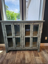 The Jofran Rustic Shores 4 Door Accent Cabinet with glass doors stands on a wooden floor by large windows.