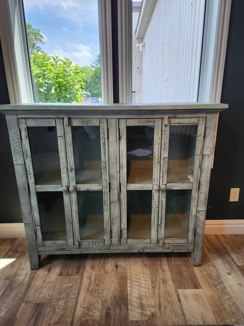 The Jofran Rustic Shores 4 Door Accent Cabinet with glass doors stands on a wooden floor by large windows.