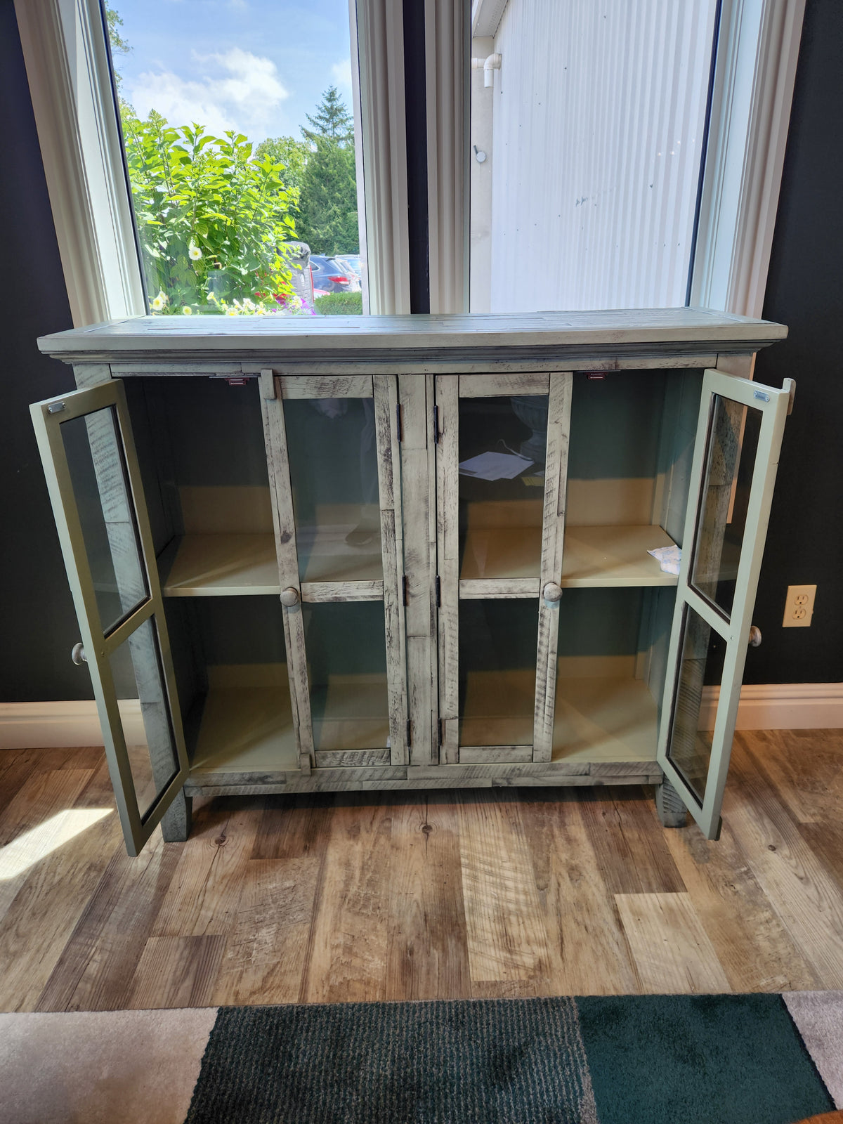 The Jofran Rustic Shores 4 Door Accent Cabinet stands open by large windows, displaying two spacious shelves.