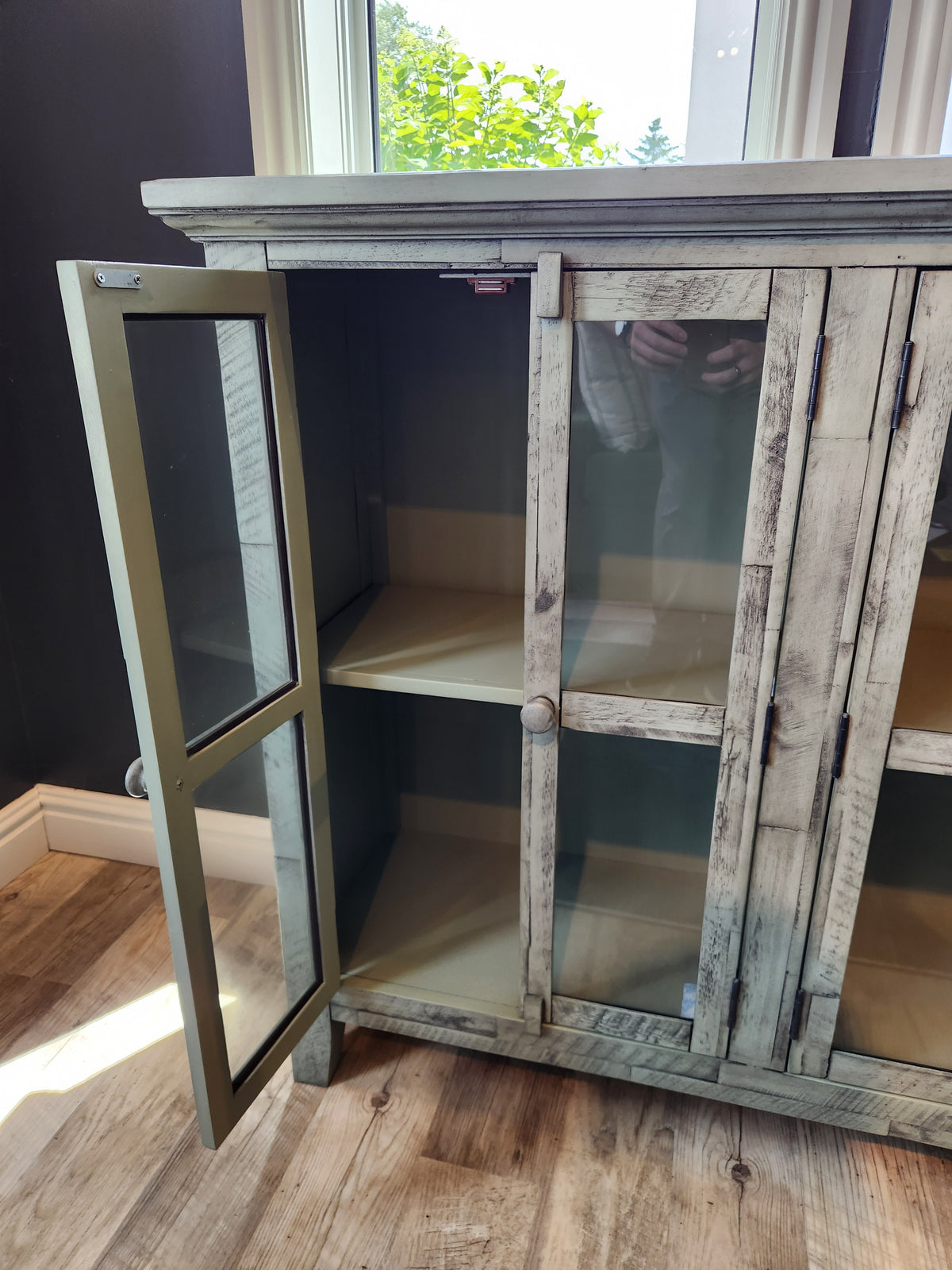 The Jofran Rustic Shores 4 Door Accent Cabinet stands open, showing empty shelves on a wooden floor beside a window.