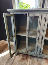 The Jofran Rustic Shores 4 Door Accent Cabinet stands open, showing empty shelves on a wooden floor beside a window.