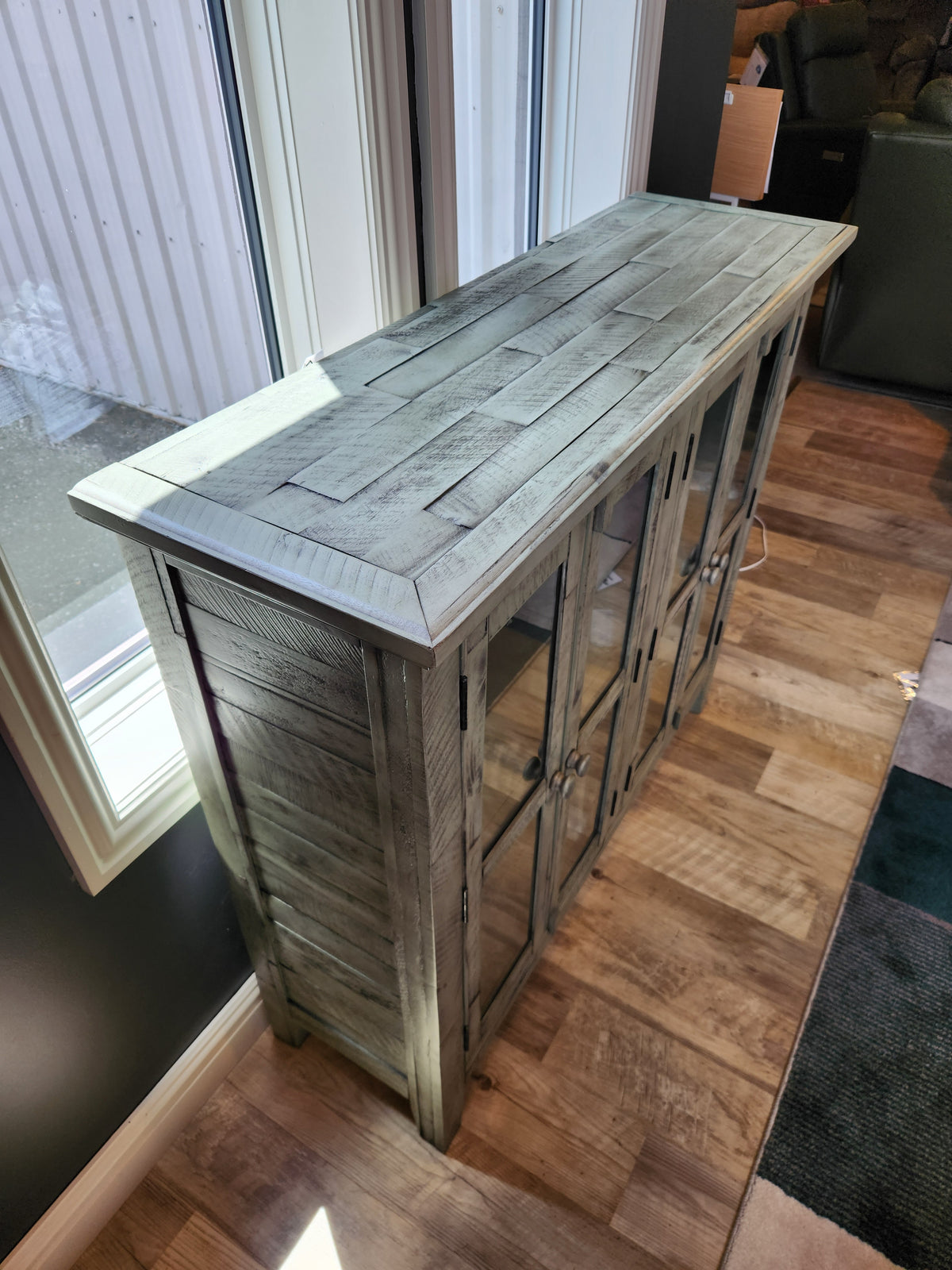 A Jofran Rustic Shores 4 Door Accent Cabinet with glass doors stands by large windows on a wood-patterned floor.