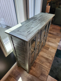 A Jofran Rustic Shores 4 Door Accent Cabinet with glass doors stands by large windows on a wood-patterned floor.