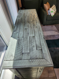 The Jofran Rustic Shores 4 Door Accent Cabinet with a distressed finish stands by a window in a sunlit room.