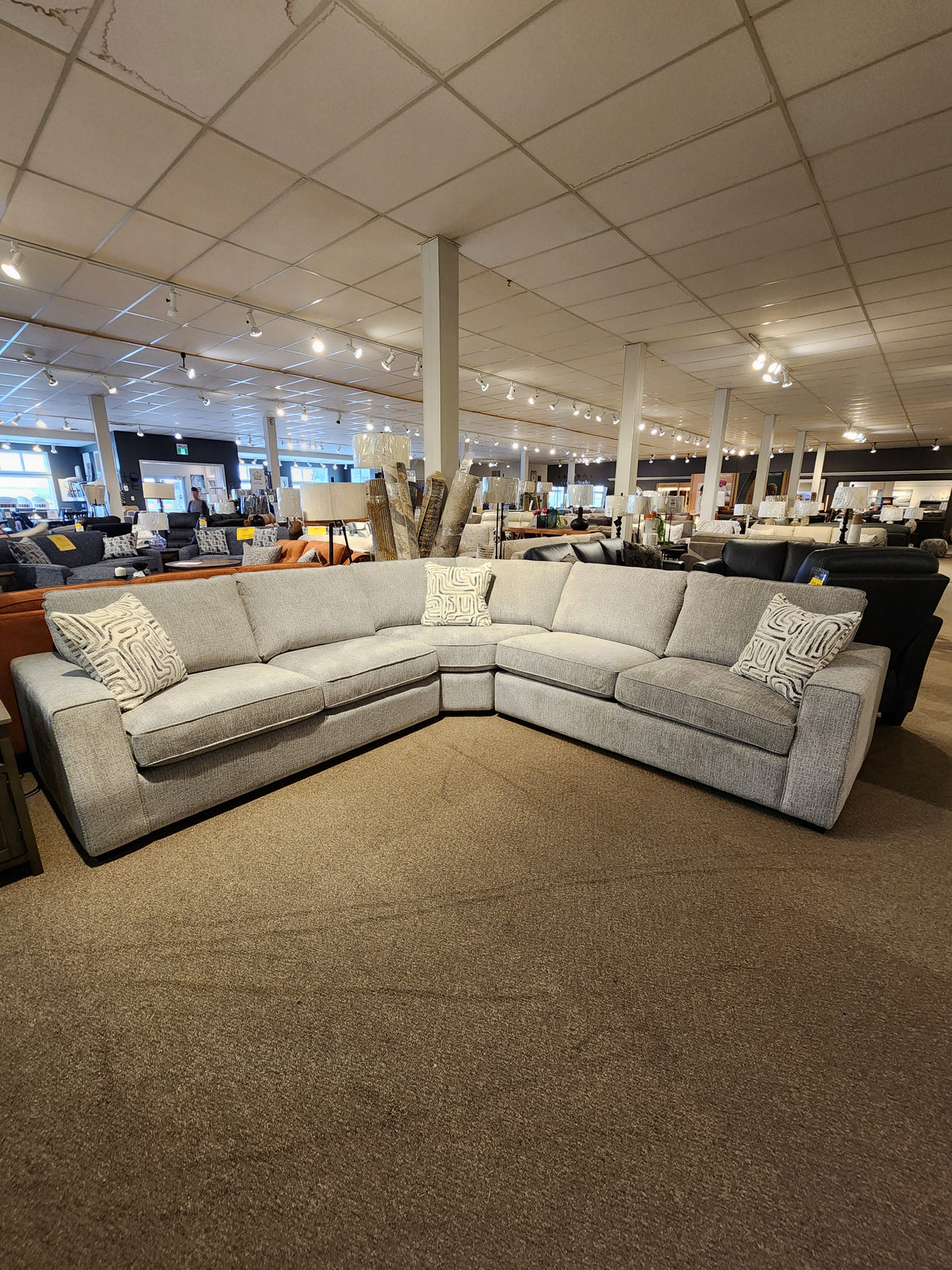 Decor-rest 2456 3-Piece Sectional in light grey with patterned pillows, shown on carpeted showroom floor under lights.