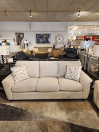A beige Superstyle 5101 Sofa with ergonomic seating and two patterned pillows is shown in a furniture showroom.