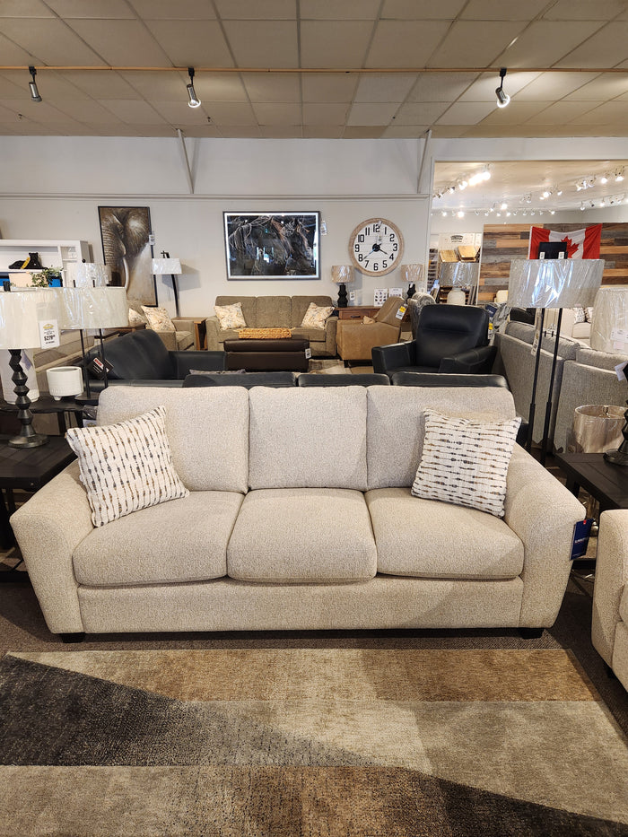 A beige Superstyle 5101 Sofa with ergonomic seating and two patterned pillows is shown in a furniture showroom.