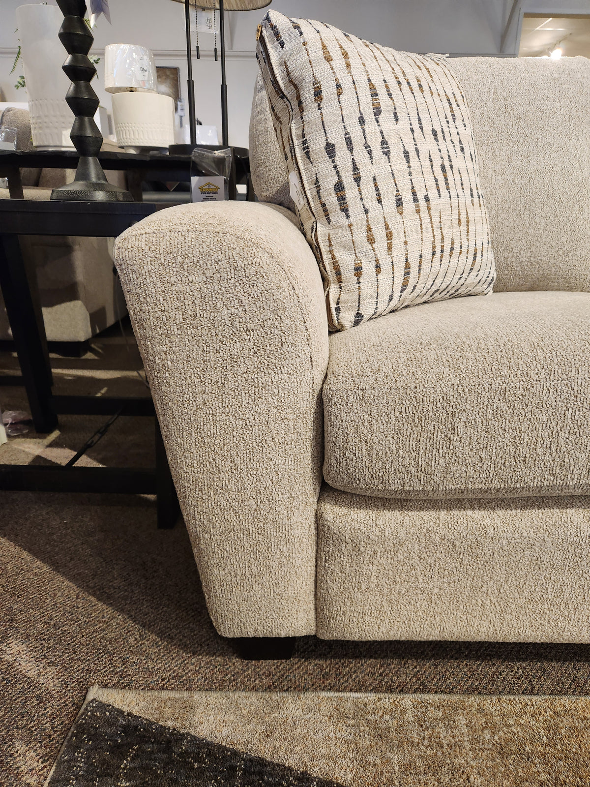 Beige Superstyle 5101 Sofa with a patterned pillow, black side table, lamp, on a brown carpeted floor.