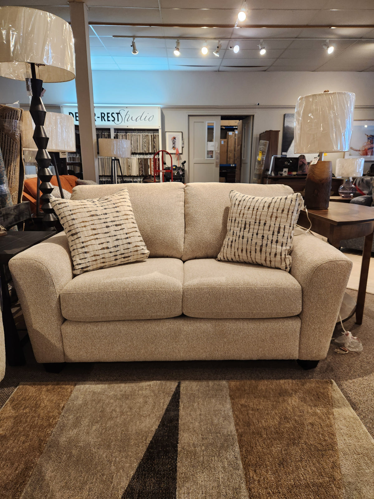 The Superstyle 5101 Loveseat comes with two patterned pillows, shown in a showroom with lamps and tables.