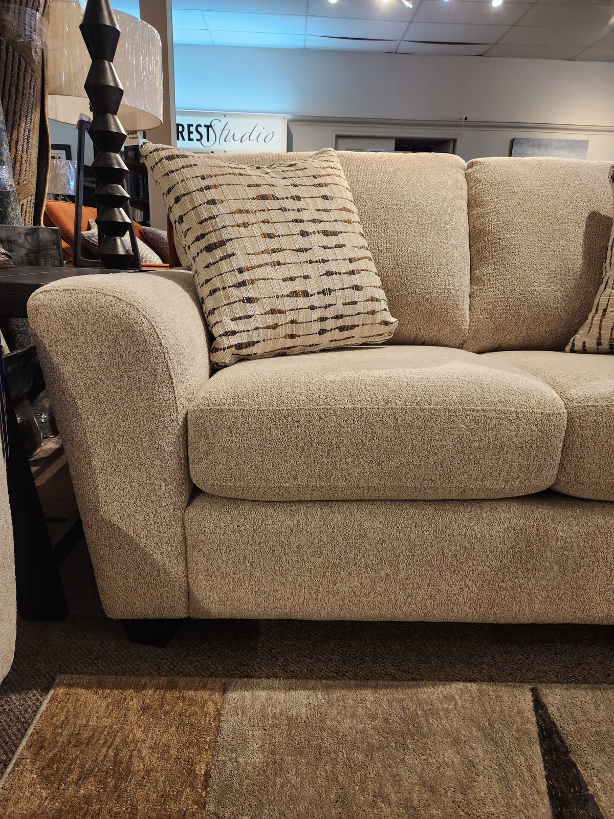 A beige Superstyle 5101 Loveseat with patterned cushions is beside a dark lamp and side table in the showroom.