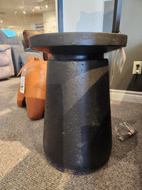 The Maru 15" Accent Table by Donald Choi, with a textured black surface, stands on carpet in a showroom.