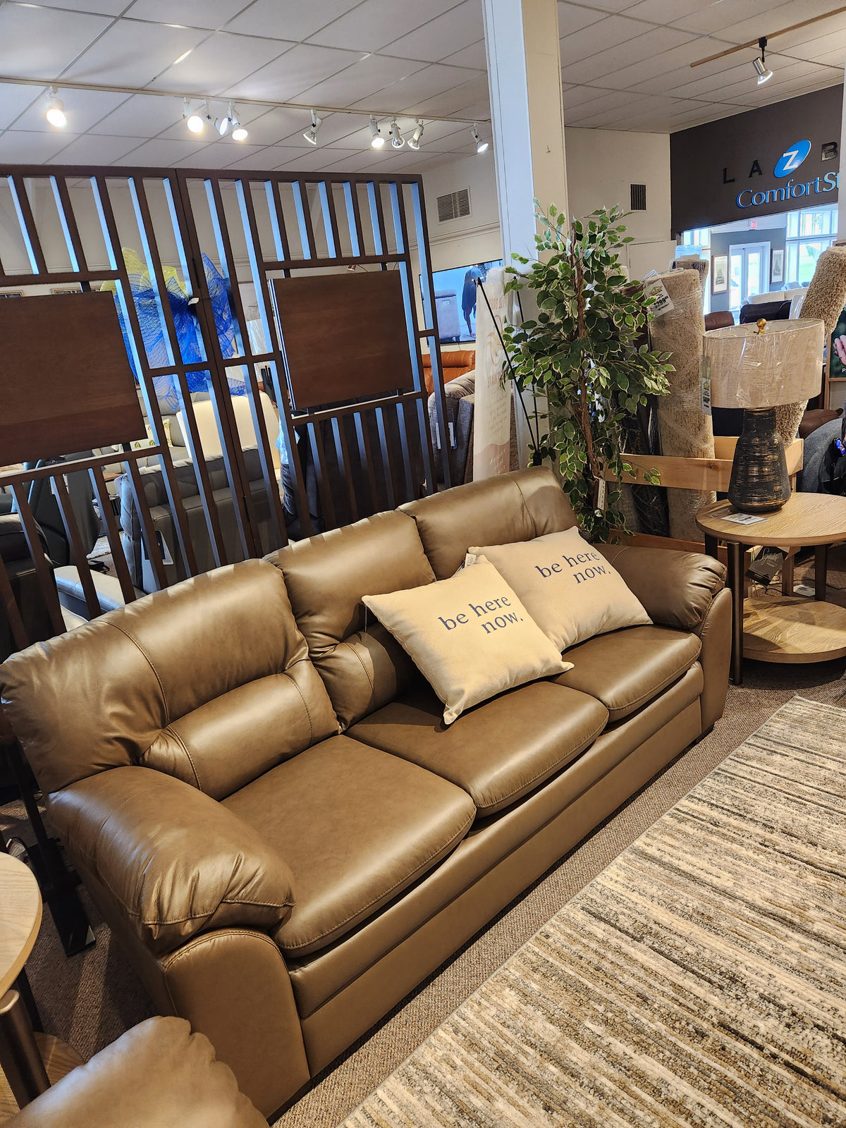 Palliser 77343 Amisk brown leather sofa with two "be here now" pillows, lounge seating, dividers, and plants.
